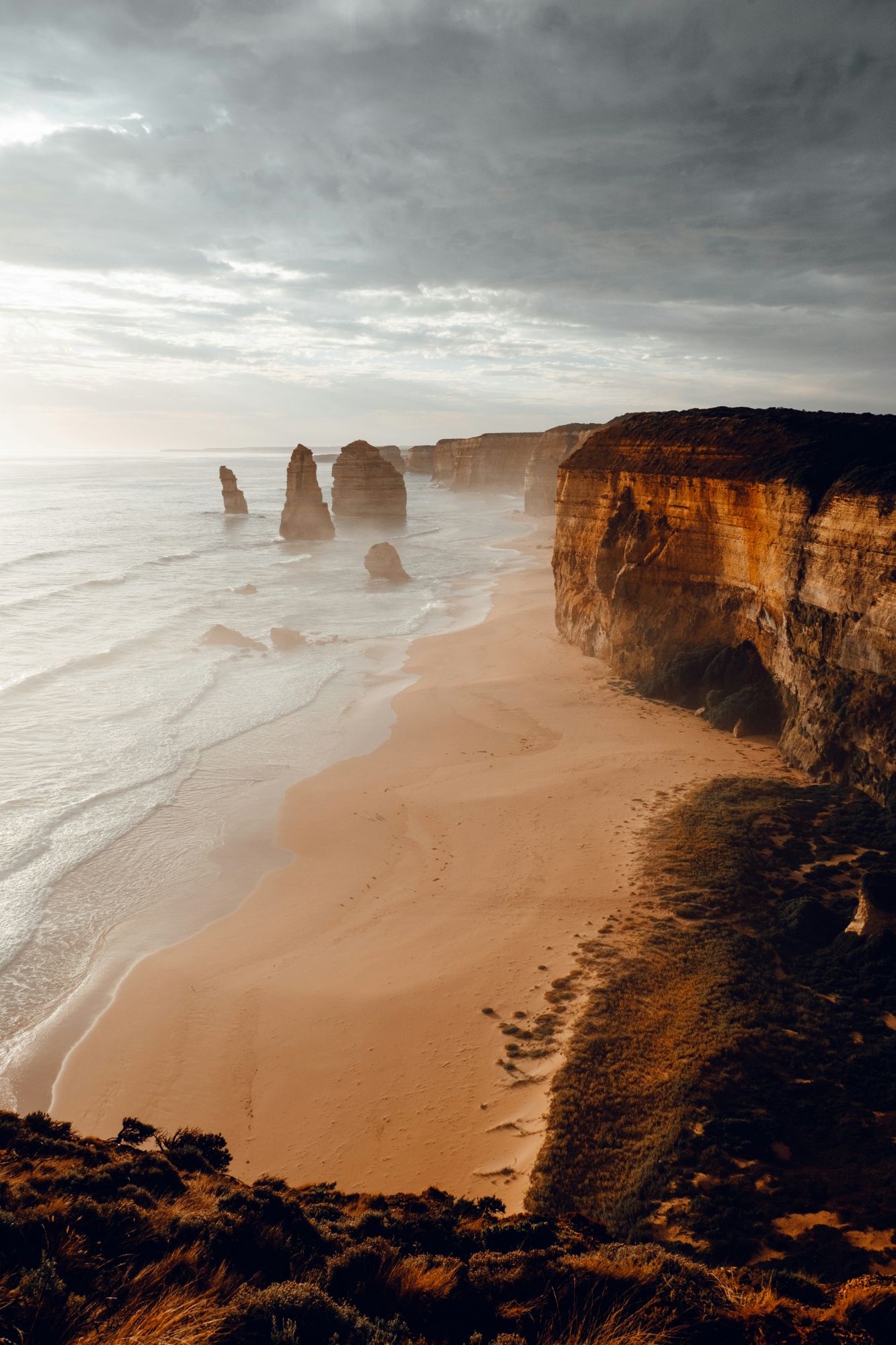 Limestone sea stacks of the Twelve Apostles beside a sandy beach under a moody overcast sky